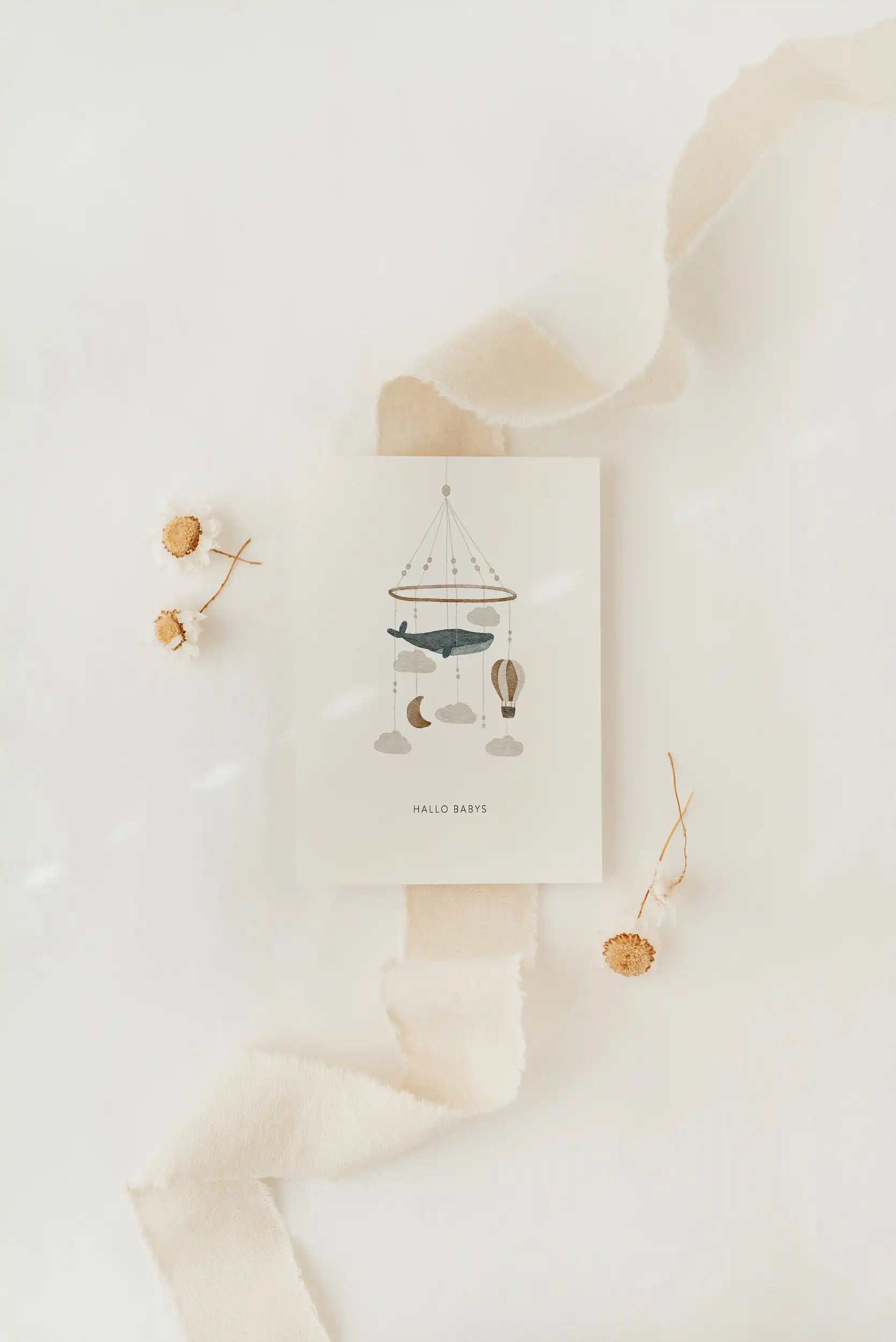Eine Postkarte von der Marke Hej Hanni mit einem Aquarellmotiv und unten in schlichtem Schriftzug ,,Hallo Baby''. Aquarellmotiv Babymobile mit wolken, mondsichel und einem Wal.